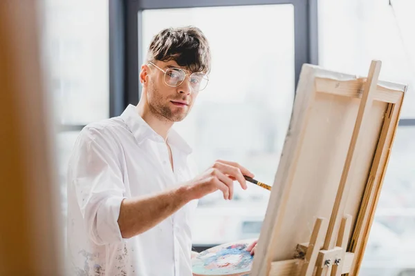 Selektiver Fokus des gutaussehenden Künstlers mit Brille, der während des Zeichnens auf Leinwand in die Kamera blickt — Stockfoto