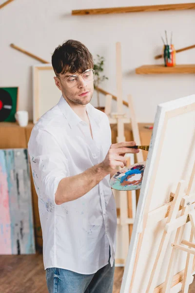 Artista guapo en camisa blanca y gafas de pintura sobre lienzo en la galería - foto de stock