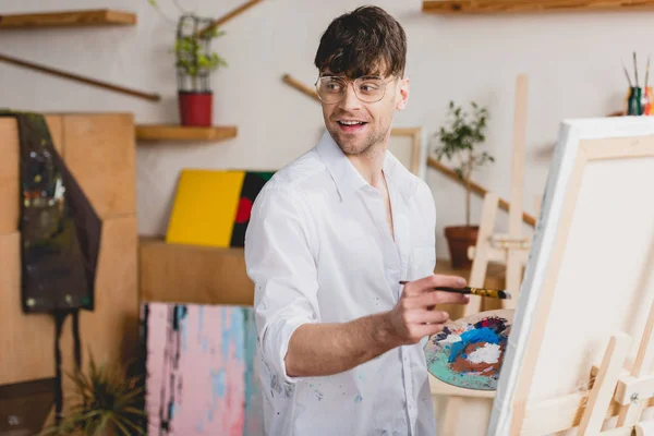 Selective focus of smiling artist in white shirt and glasses painting on canvas — Stock Photo