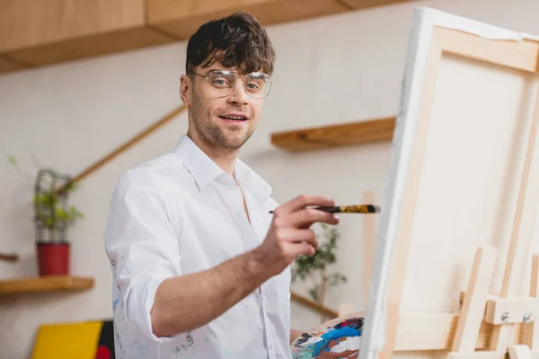 Selective focus of smiling artist in white shirt and glasses painting on canvas and looking at camera — Stock Photo