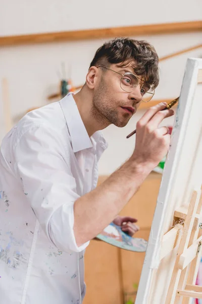 Handsome artist in white shirt and glasses painting on canvas — Stock Photo