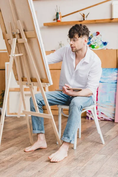 Handsome artist in white shirt and glasses painting on canvas while sitting on chair in painting studio — Stock Photo