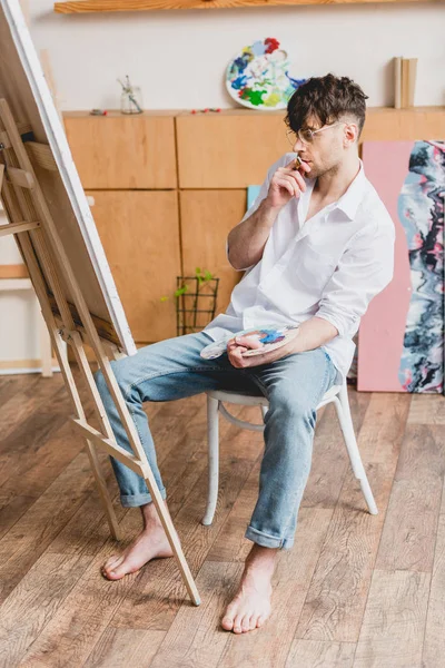Bello artista in camicia bianca e jeans blu guardando tela su cavalletto — Stock Photo
