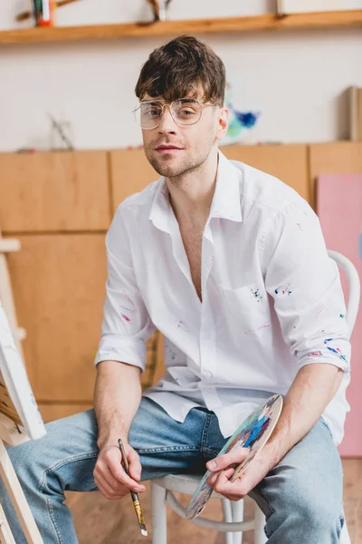Handsome artist holding palette and paintbrush while sitting and looking at camera — Stock Photo