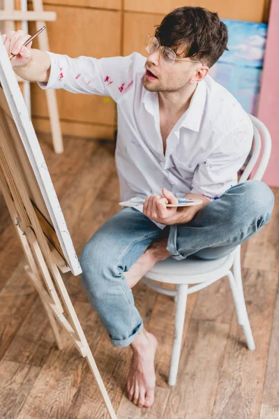 Selektiver Fokus des gut aussehenden Künstlers auf Leinwand, während er auf einem Stuhl im Malatelier sitzt — Stockfoto