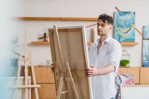 Selective focus of handsome artist looking at camera while standing at easel with canvas — Stock Photo