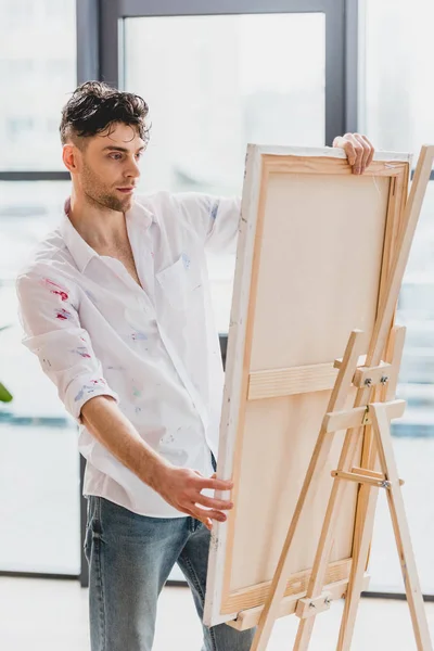 Gutaussehender Künstler im weißen Hemd fixiert Leinwand auf Staffelei im Malatelier — Stockfoto