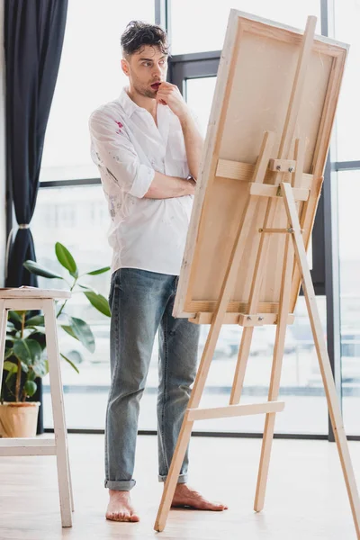 Thoughtful artist in white shirt and blue jeans standing at easel in gallery — Stock Photo