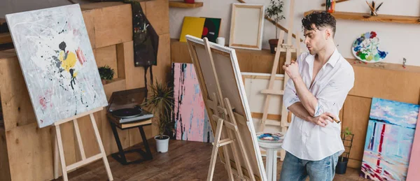 Panoramic shot of handsome artist standing at easel in painting studio — Stock Photo