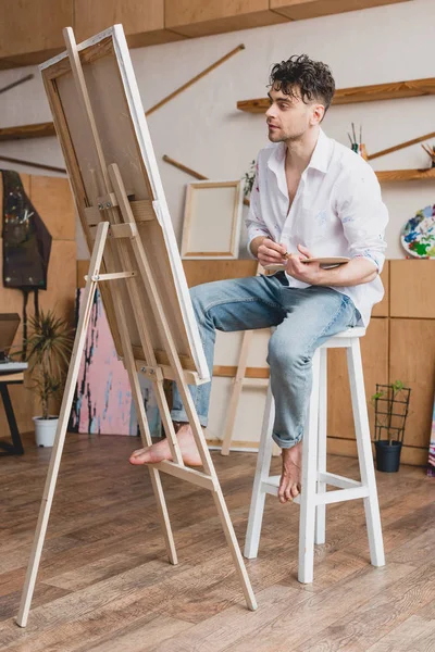 Handsome artist in white shirt and blue jeans sitting at easel in painting studio — Stock Photo
