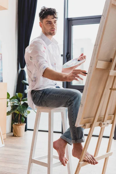 Handsome artist in white shirt and blue jeans painting on canvas in gallery — Stock Photo