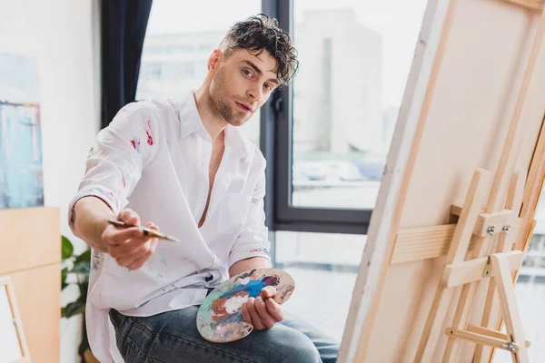 Good-looking artist with palette and paintbrush looking at camera — Stock Photo