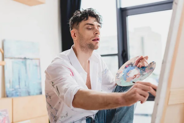 Selective focus of handsome artist painting on canvas with paintbrush — Stock Photo