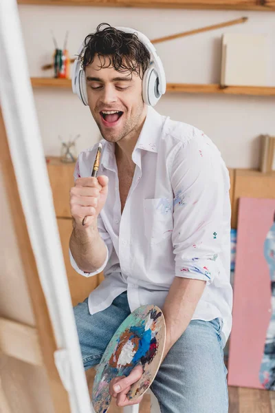 Selective focus cheerful artist in headphones, singing with paintbrush while holding palette — Stock Photo