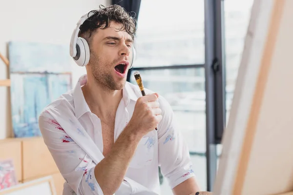 Selektiver Fokus eines gut aussehenden Künstlers mit Kopfhörer, der mit Pinsel singt — Stockfoto