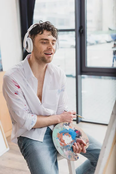 Handsome smiling artist in headphones listening music while holding palette and paintbrush — Stock Photo