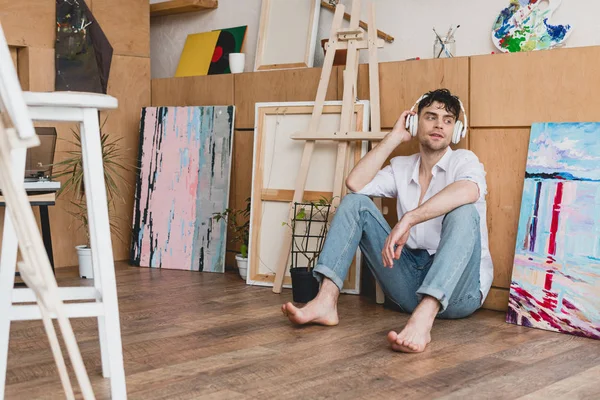 Pensive artist in headphones, listening music while sitting on floor in painting studio — Stock Photo