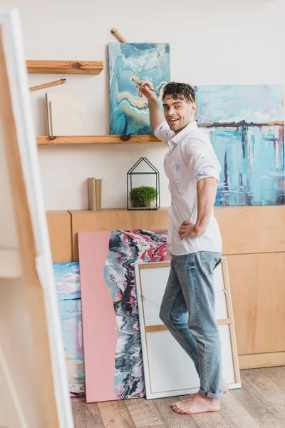 Selective focus of handsome artist standing in painting studio and looking at camera — Stock Photo