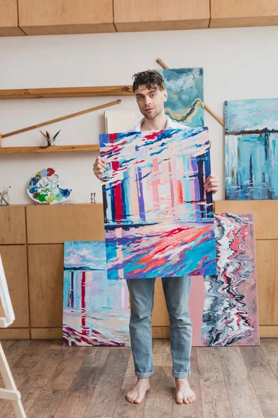 Handsome artist holding colorful abstract painting and looking at camera — Stock Photo