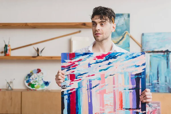 Artista de boa aparência segurando pintura abstrata multicolorida e olhando para longe — Fotografia de Stock