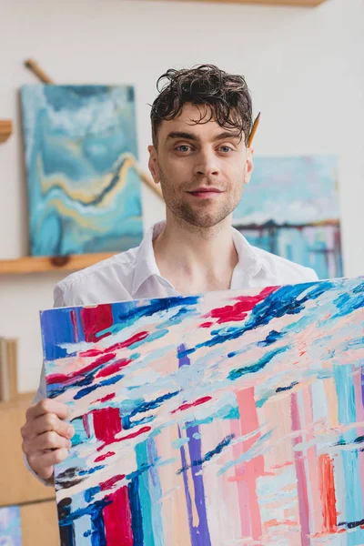 Красивый художник держит многоцветную абстрактную живопись и смотрит в камеру — стоковое фото