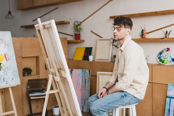 Handsome artist in eyeglasses sitting near easel with canvas in painting studio — Stock Photo