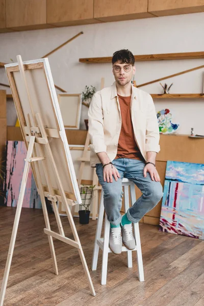 Gutaussehender Künstler mit Brille sitzt auf einem Hochstuhl in der Galerie und blickt in die Kamera — Stockfoto