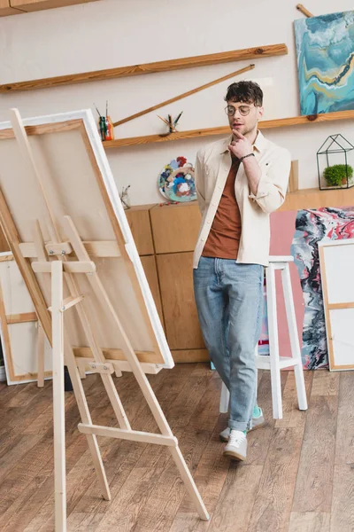 Schöner Künstler in blauen Jeans und rosa Hemd steht im Malatelier und schaut auf Staffelei mit Leinwand — Stockfoto