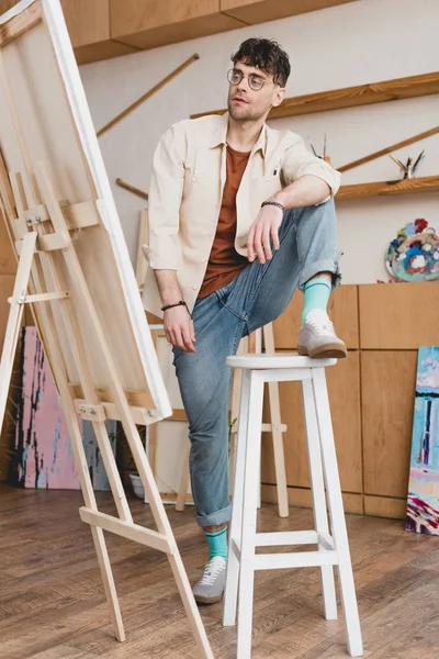 Beautiful artist standing with foot on chair and looking at easel with canvas — стоковое фото