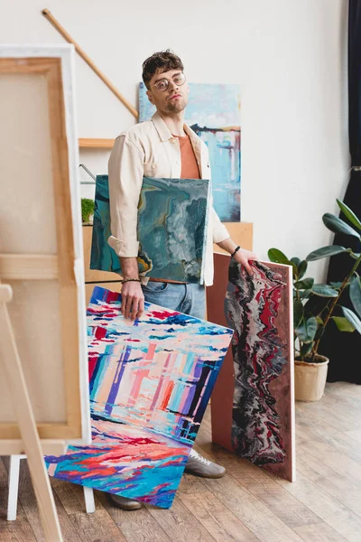 Serious artist in eyeglasses holding colorful paintings and looking at camera — Stock Photo