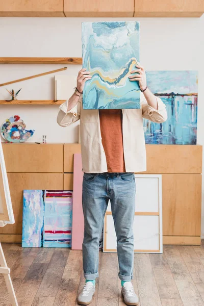 Artista de pé no estúdio de pintura e escondendo o rosto por trás da pintura abstrata colorida — Fotografia de Stock