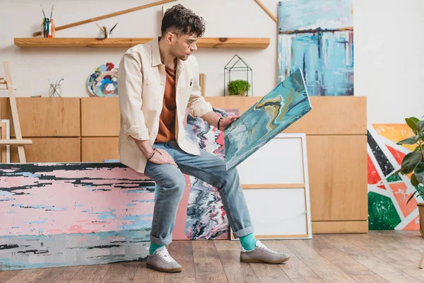 Handsome artist in pink shirt and blue jeans looking at colorful abstract painting — Stock Photo