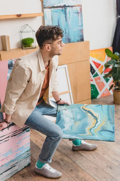 Artista guapo en camisa rosa y vaqueros azules sosteniendo pintura abstracta colorida y mirando hacia otro lado - foto de stock