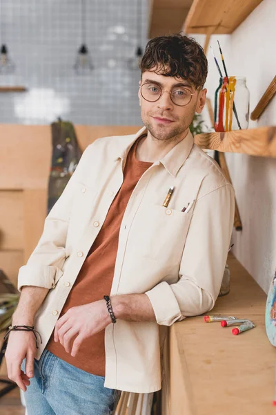 Selektiver Fokus des gutaussehenden Künstlers mit Brille, der im Malatelier steht und in die Kamera blickt — Stockfoto