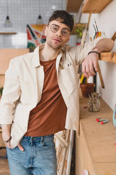 Selective focus of handsome artist  in eyeglasses leaning on wooden shelve in painting studio — Stock Photo