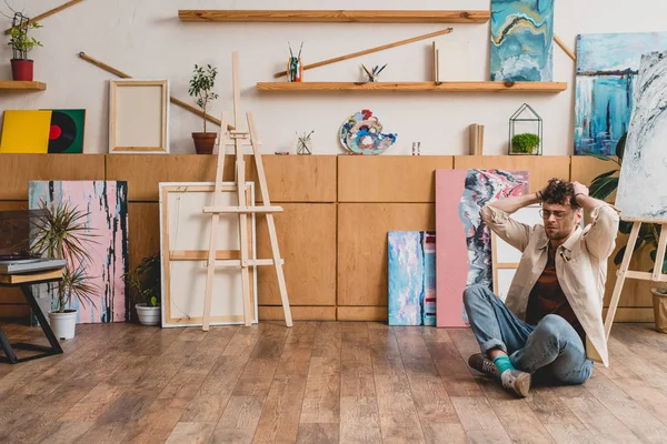 Artista sentado en el suelo con las piernas cruzadas y las manos en la cabeza en el estudio de pintura - foto de stock