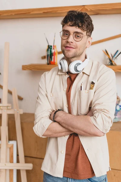 Beau artiste avec écouteurs sur le cou et les bras croisés regardant la caméra — Photo de stock