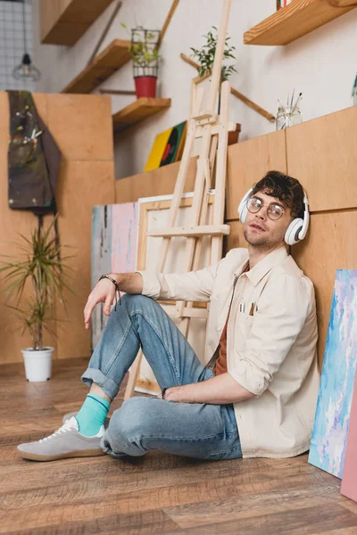 Artista pensativo en auriculares sentados en el suelo y escuchando música en el estudio de pintura - foto de stock