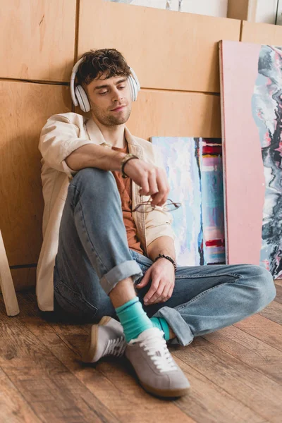 Selective focus of handsome artist sitting on wooden floor and listening music in headphones — Stock Photo