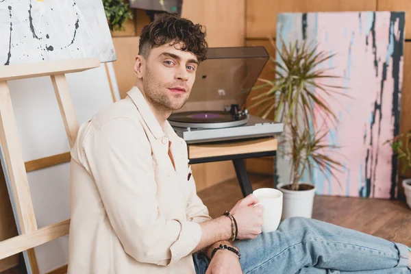 Artista guapo sosteniendo taza de café mientras está sentado cerca de vinilo tocadiscos - foto de stock