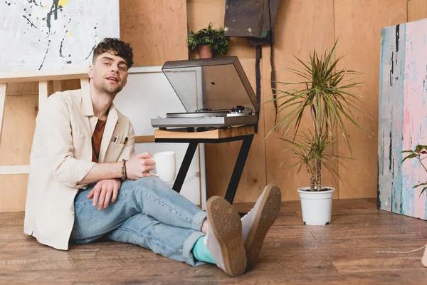 Beau artiste tenant tasse de café et regardant la caméra tout en étant assis près du lecteur de disque vinyle — Photo de stock