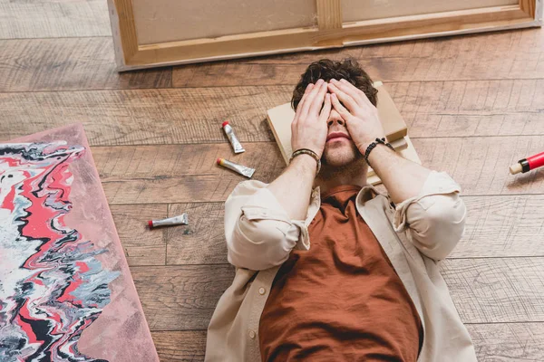 Overhead view of exhausted artist lying on wooden floor and holding hands on face — Stock Photo