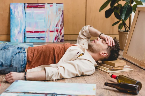 Selective focus of exhausted artist lying on floor and holding hand on head — Stock Photo