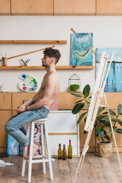 Handsome half-naked artist in blue jeans sitting on high chair in painting studio — Stock Photo