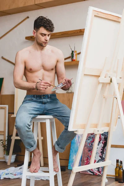 Schöner halbnackter Künstler mischt Farben auf Palette, während er auf einem Hochstuhl in der Galerie sitzt — Stockfoto