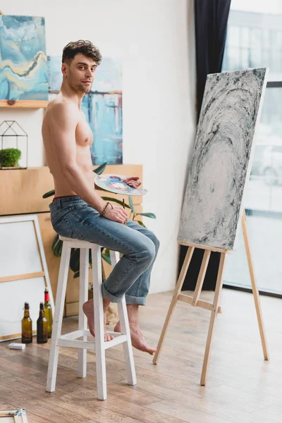 Good-looking half-naked artist looking at camera while sitting on high chair near easel with painting — Stock Photo