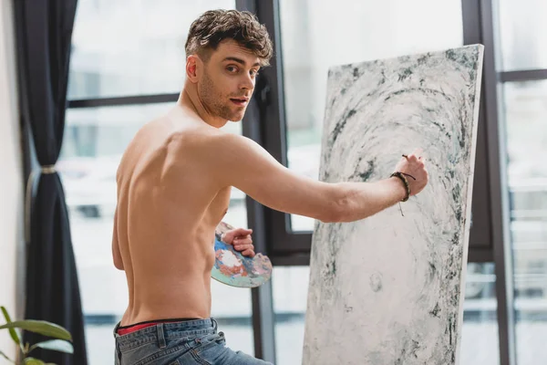 Guapo artista semidesnudo mirando a la cámara mientras pinta cuadro - foto de stock