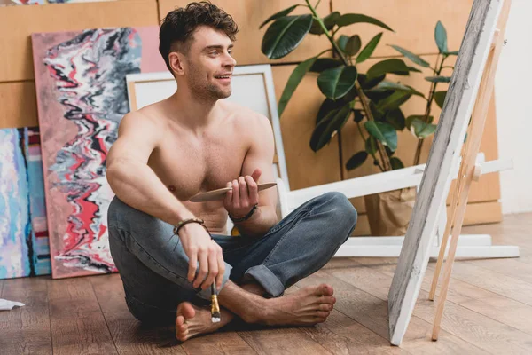 Smiling half-naked artist sitting with crossed legs on floor in painting studio — Stock Photo