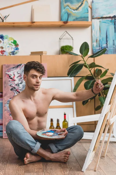 Handsome half-naked artist holding palette and painting picture on canvas — Stock Photo