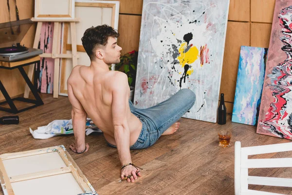 Handsome half-naked artist sitting on floor in front of canvas with picture — Stock Photo